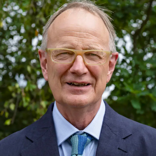 A white man in his late sixties is smiling at the camera. He is wearing yellow trendy glasses and a blue suit, with a crisp white shirt.