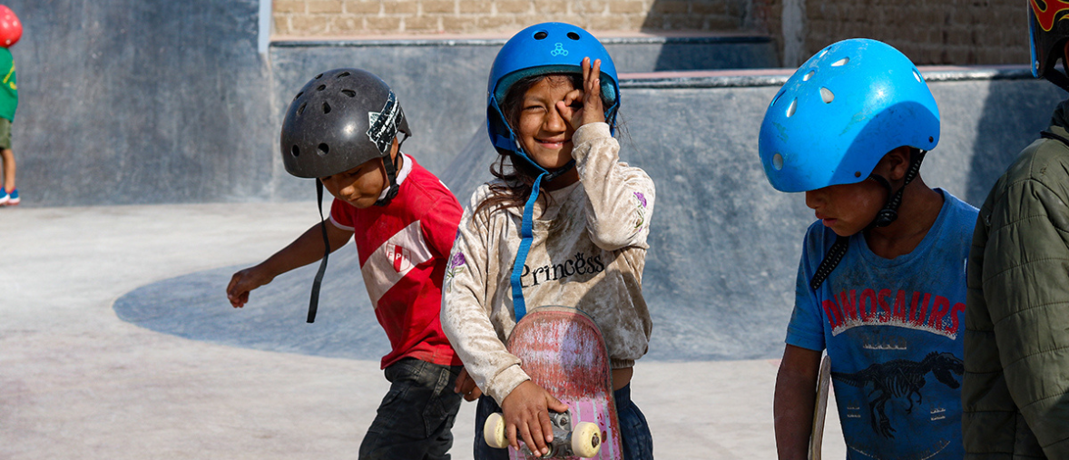 Three children in brightly coloured skate helmets, and skateboards are playing in a concrete skatepark. One child with long hair is smiling and looking at the camera with her hands in a circle around one eye, and holding her skateboard in the other.