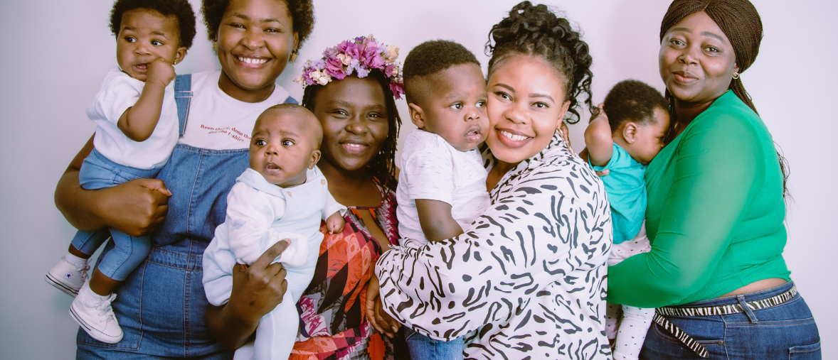 Four Black women and their babies are all cuddling and standing together, smiling widely, on a professional photography background. The babies are all around 0-2 years old and incredibly cute.
