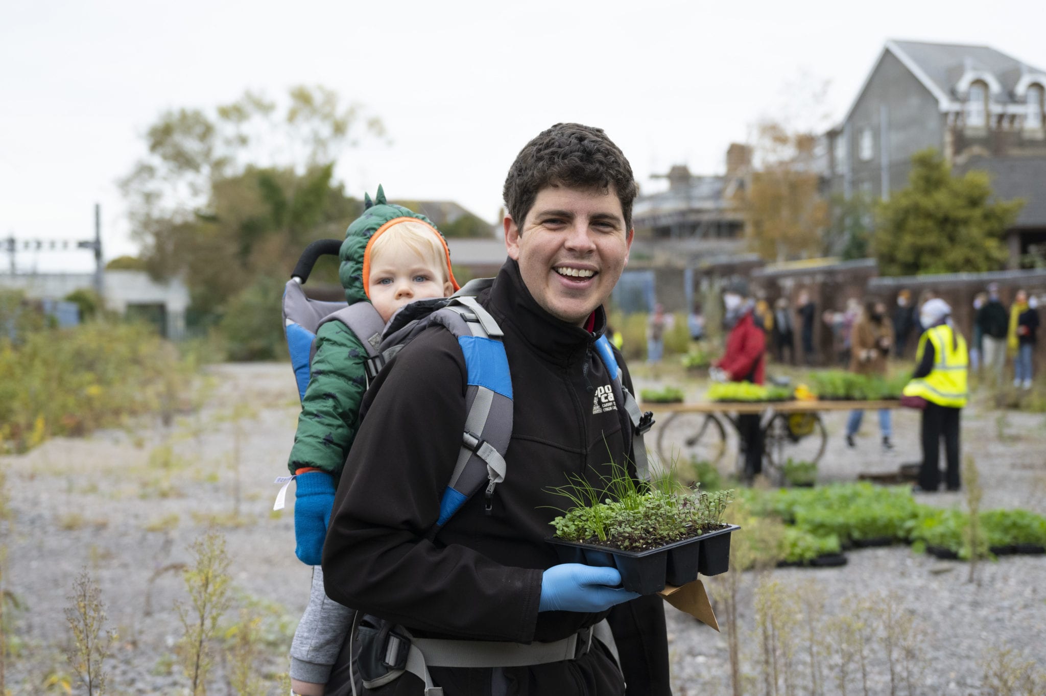 A man and his baby carrying plants, dressed in winter clothing. The baby is in his backpack.