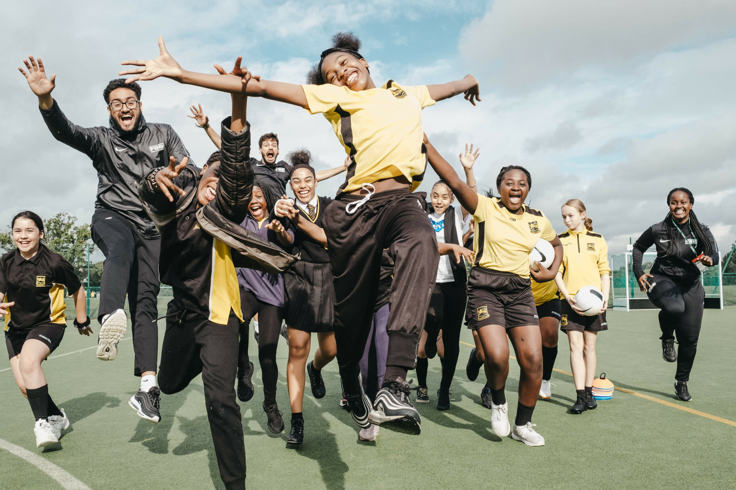 A group of around 12 children and teens, jumping, spreading their arms in joy and smiling at the camera. They are stood on a football field and wearing yellow football kits or school uniform.